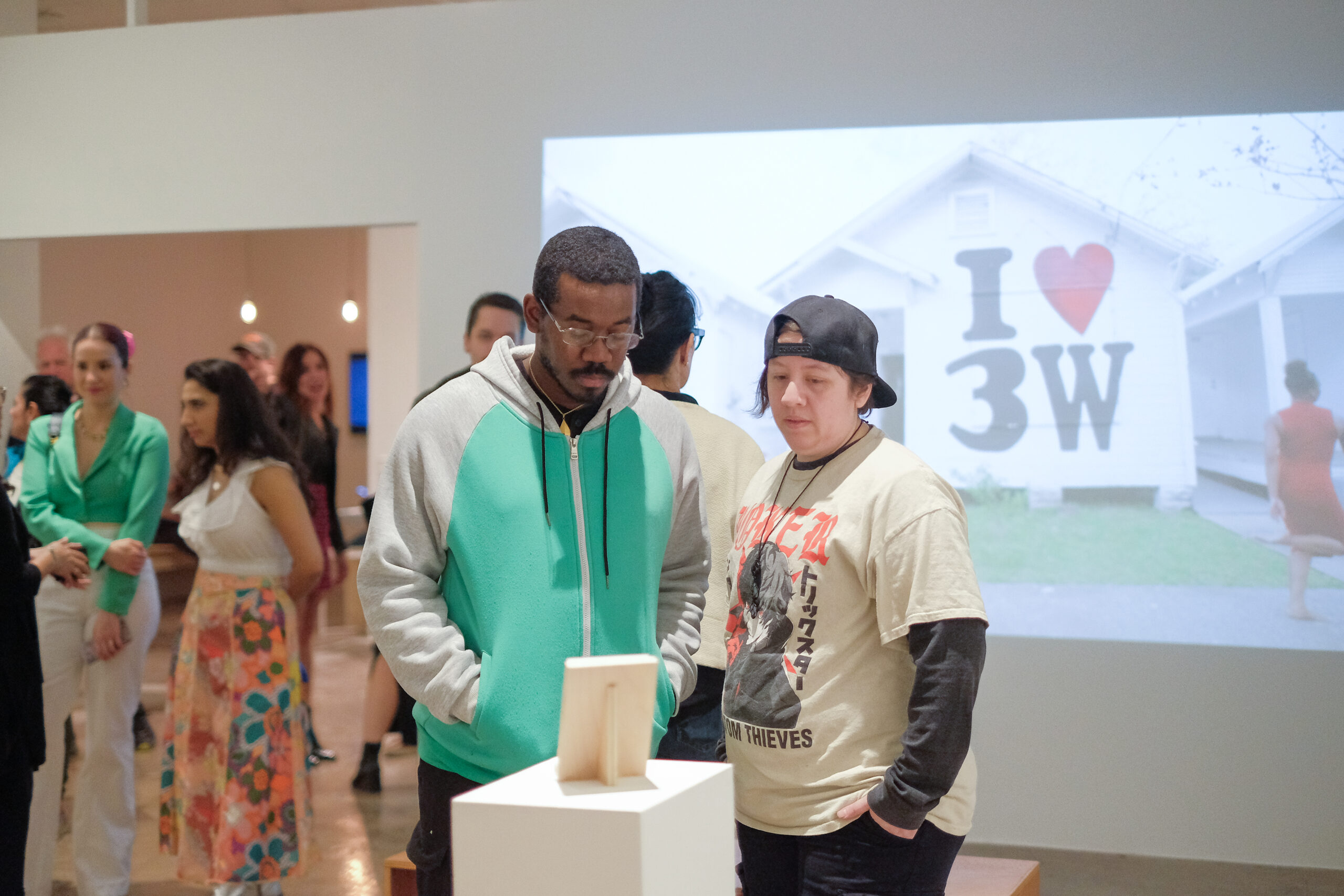 Two people in a gallery looking at a small frame placed on a pedestal. A projection is on the wall in the background, showing a White House with I (heart) 3W on it.