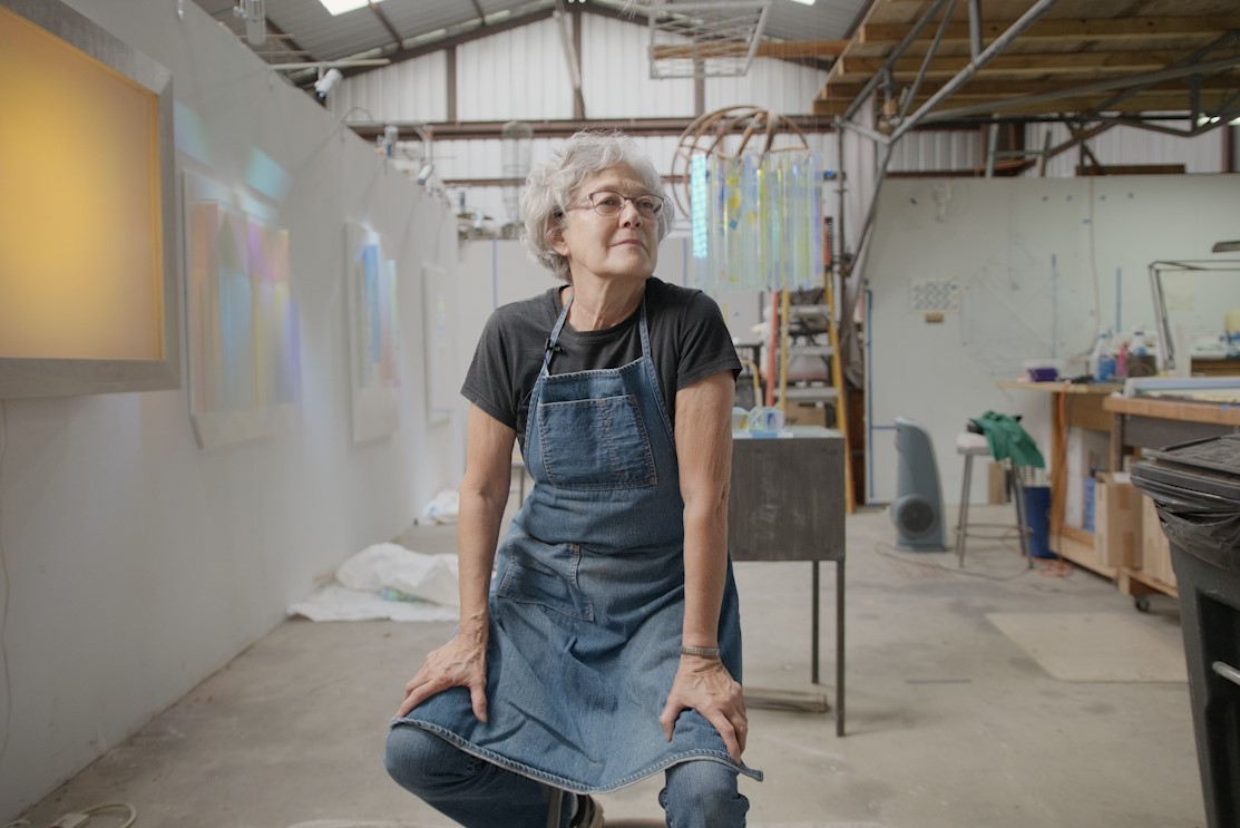 Woman artist Cathy Cunningham-Little in an apron and black tshirt sitting on a stool in the middle of her studio space.