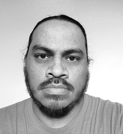 Joe Harjo, a brown, Native American man of the Muskogee nation, stares directly into the camera, looking at the viewer of the photograph. The photograph is in black and white with a solid light gray background. Joe has black facial hair, including a short beard and mustache. He also has black hair.