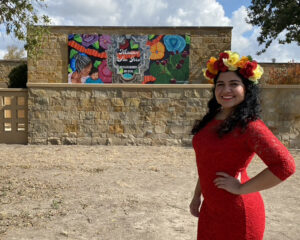 Image of artist Sandra Gonzalez wearing a red dress and a flower crown in the foreground of her mural at Mission San Jose.