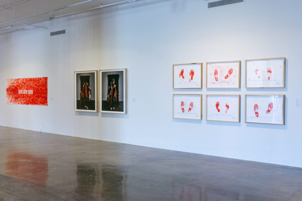 Image of three artworks on a wall, two of which feature a man dressed in modern clothing meant to allude to the Virgin Mary. And six framed works on paper, featuring red footprints on each.