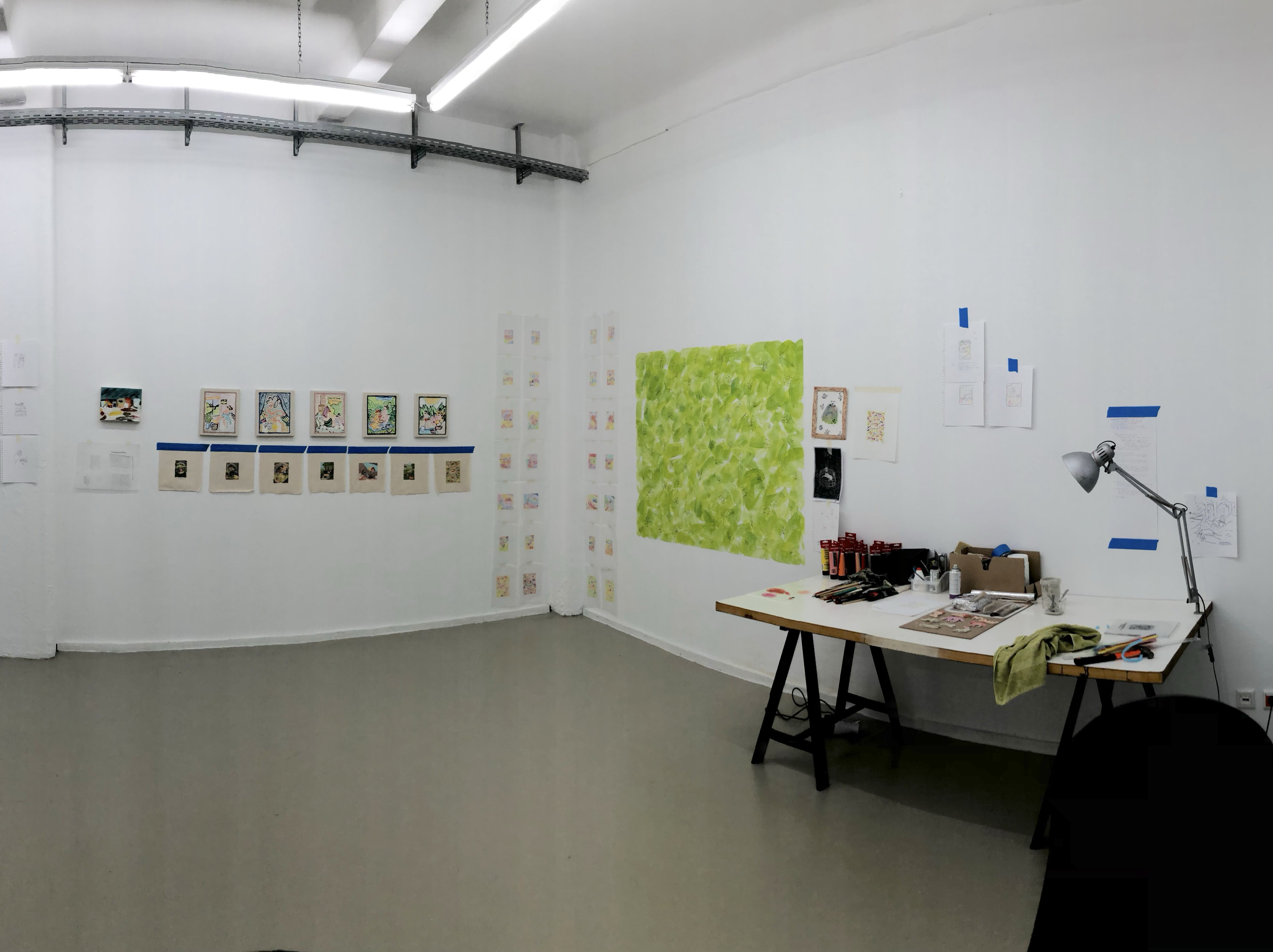 Photograph shows a 180 degree view of an artist studio. The right side of the frame there are three large windows one of which is open. The frames a pulled open and there is a view of a 6 story building out it. In the center of the frame is an artists work table with a lamp and materials and tools. On the walls there is one large bright green painting, a large figurative sketch and numerous small studies for paintings and drawings on paper, taped up on the wall with masking tape.
