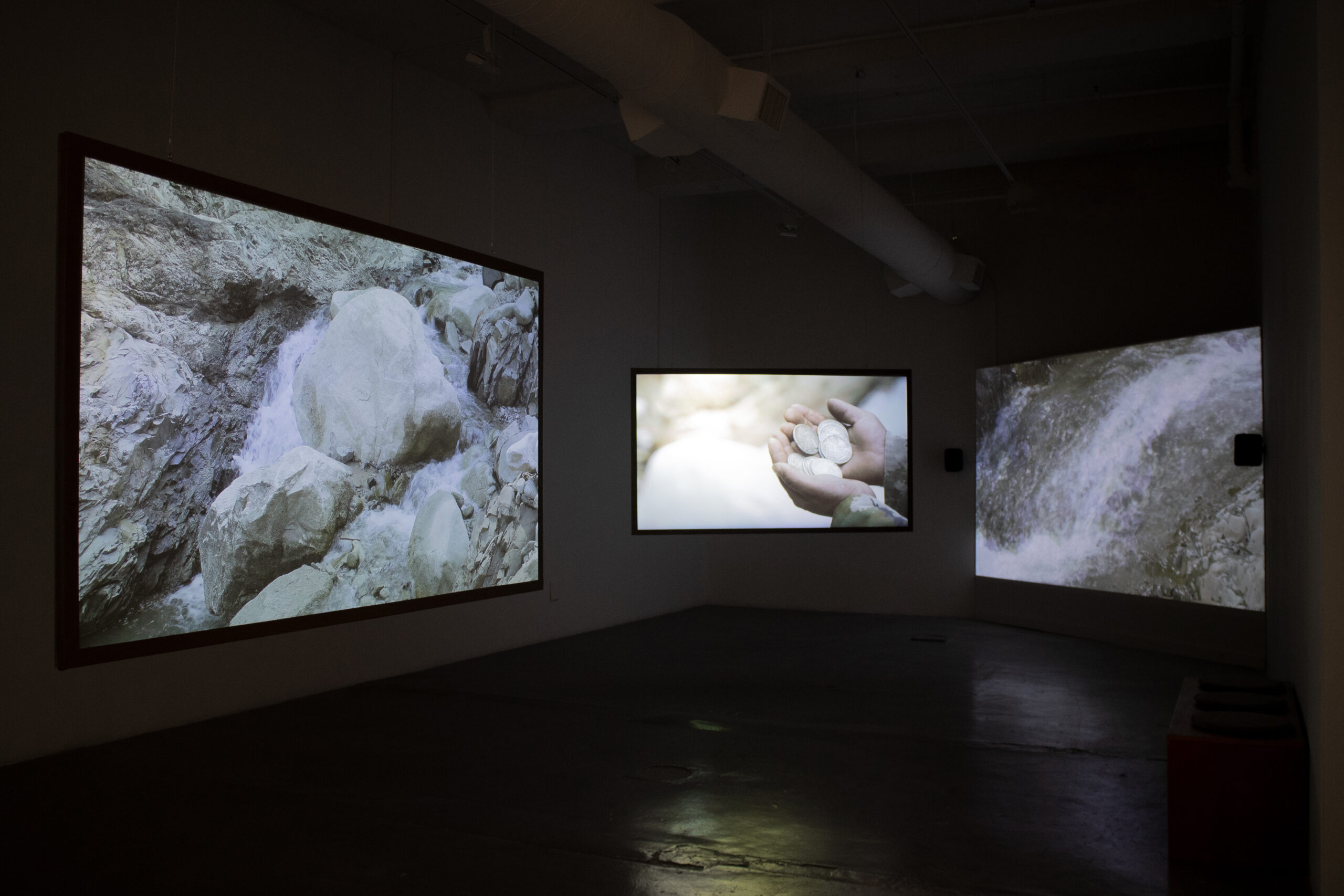 A dark gallery with three screens. From left to right, a stream over rocks, two hands cupped together holding silver coins, and another stream.
