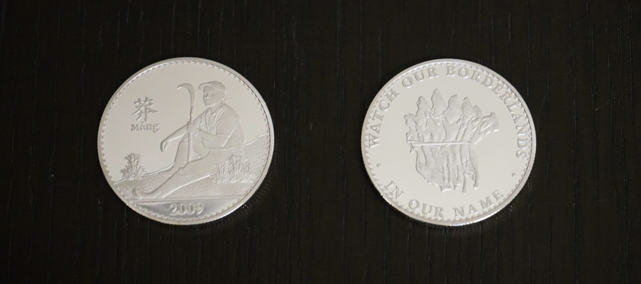 Two silver coins place side by side on black velvet. The left coin depicts a man sitting, holding a sickle with inscriptions of the year 2009 and Mang, an ethnic group of people who live along the China-Vietman border.