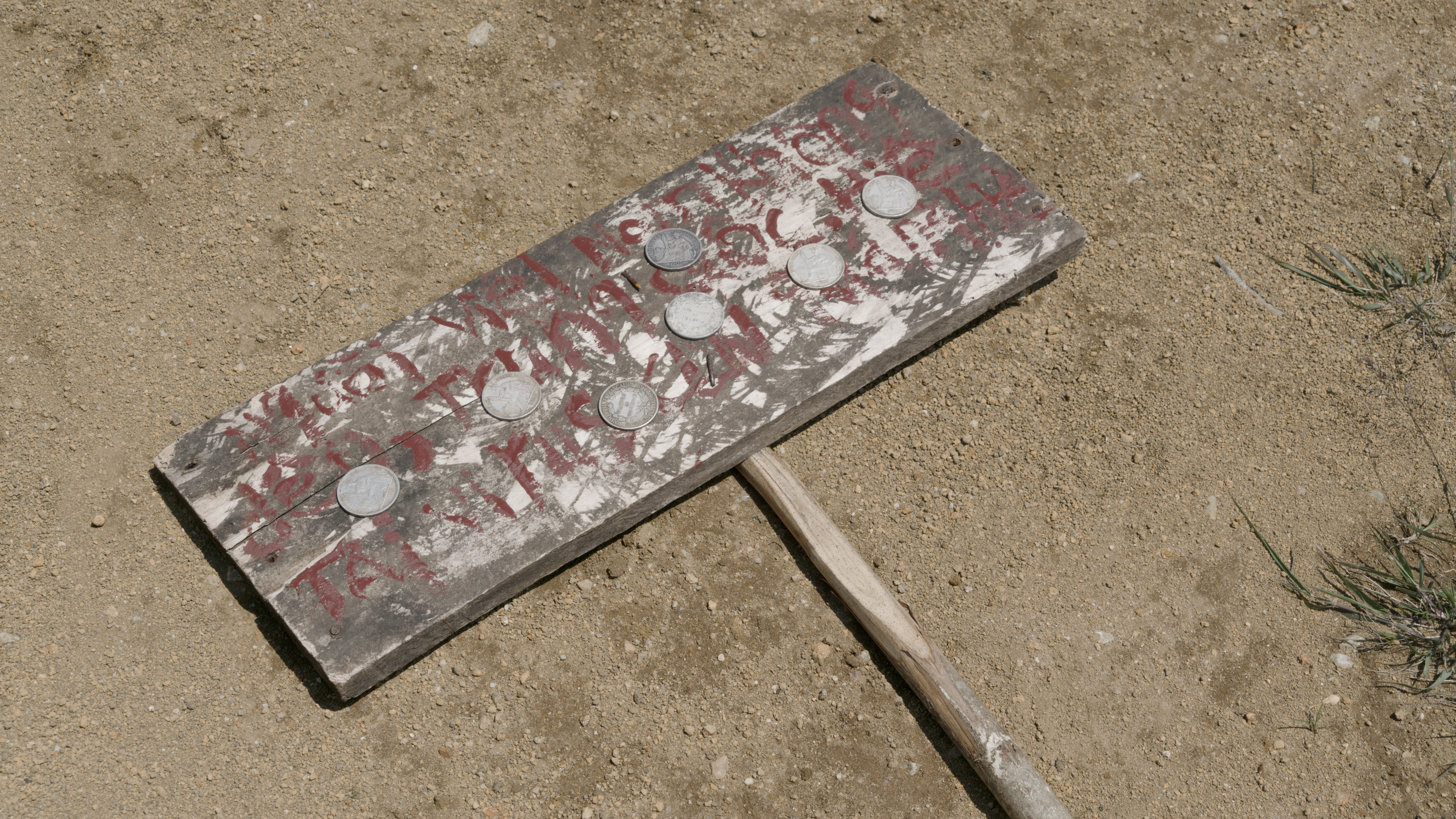 A wooden sign with red text sits on course dirt. & silver coins are places on the wooden sign, which has red text.
